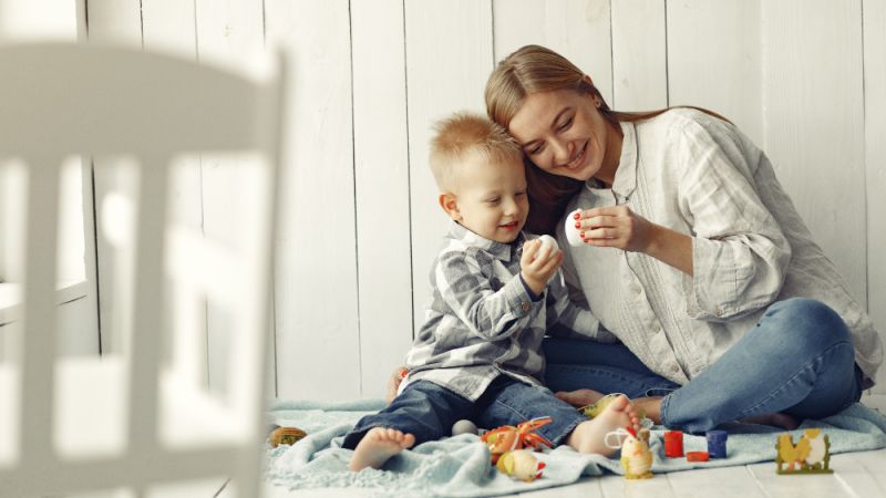Mother and young child sitting on a blanket indoors, smiling while holding decorative eggs and playing with toys in a bright, white wooden room.